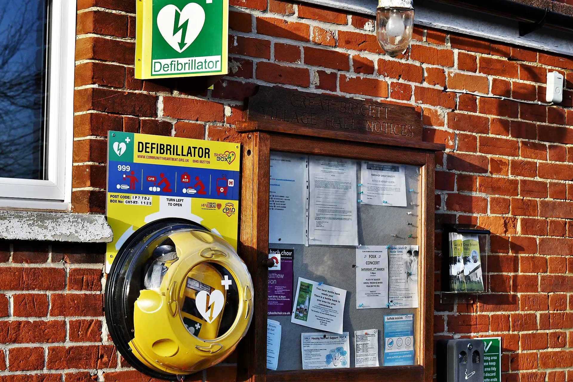 A community hall notice board with posted notices on a brick wall, beside a defibrillator.